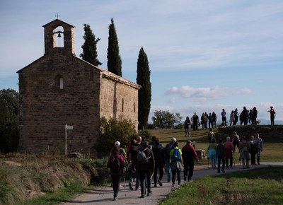 Imatge cedida per Laia Ausió. Persones caminant per un sender local i una ermita a la banda esquerra de la foto..
