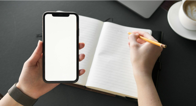 Una mà subjecta un mòbil amb la pantalla en blanc i l'altra escriu sobre una llibreta en blanc..