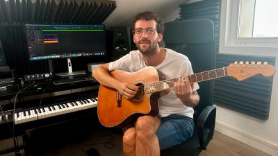 Imatge de Gil Grau assegut davant un piano, subjectant una guitarra en una habitació.